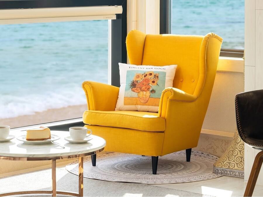 Ein gelber Sessel mit Kissen, das ein Gemälde von Vincent van Gogh zeigt, ein kleiner runder Tisch mit zwei Tassen und einem Stück Kuchen, und Meerblick durch ein Fenster.
