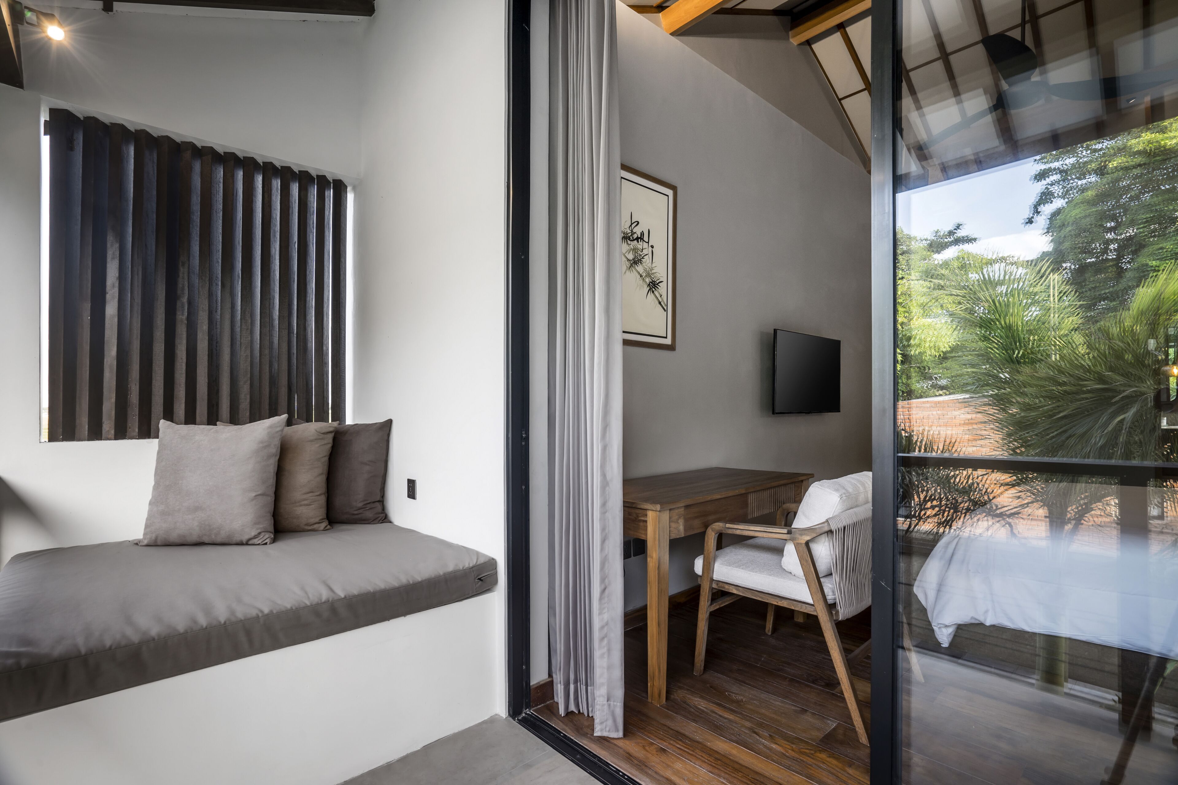 A modern room with a bed, a wooden desk, and a chair. There is a window with a view of greenery outside.