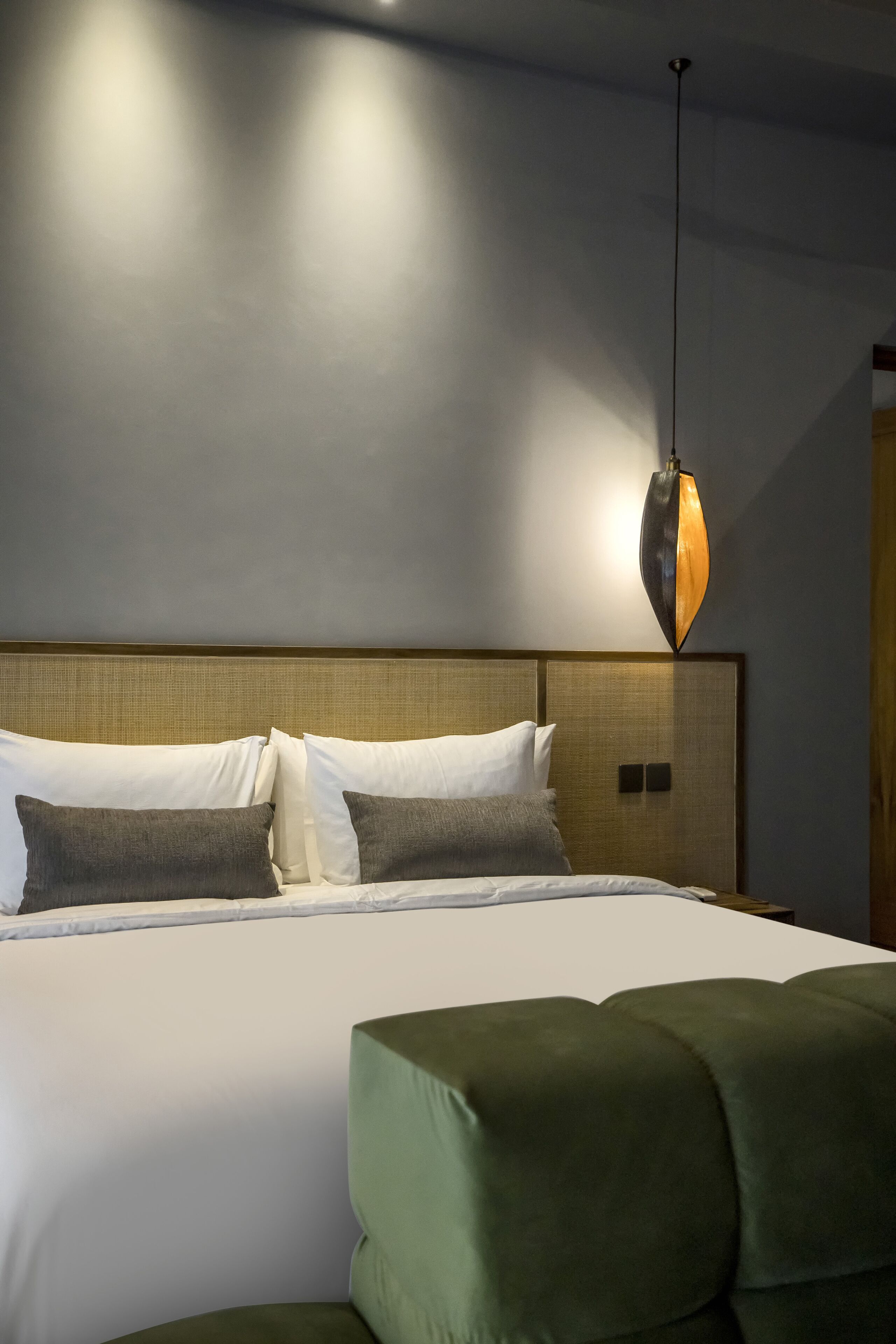 A modern hotel room with a large bed, a green upholstered headboard, and a unique pendant light.