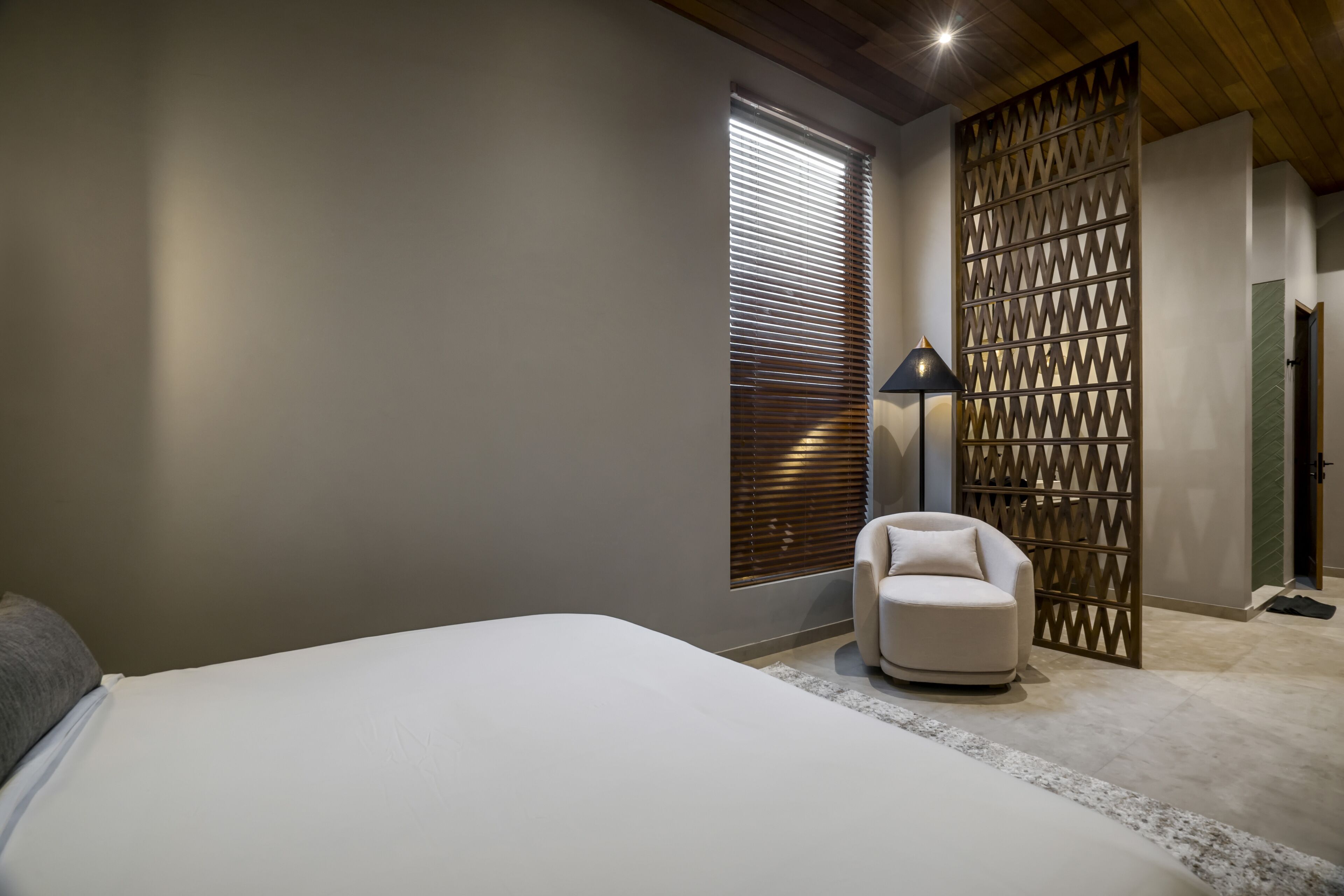 Un dormitorio con cama, una silla, ventana con persianas, lámpara y un separador decorativo.