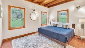 A bedroom with a wooden ceiling, large windows with wooden frames, a bed with a blue bedspread, and a patterned rug. - Pet-Friendly Cabin in Marietta w/ Porch Swings (Cleveland)