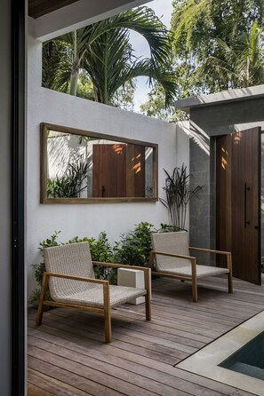 A modern outdoor patio with two wicker chairs, a wooden deck, a pool, and a large mirror reflecting the surrounding greenery. - Villa Pereh by Alfred in Bali (Canggu)