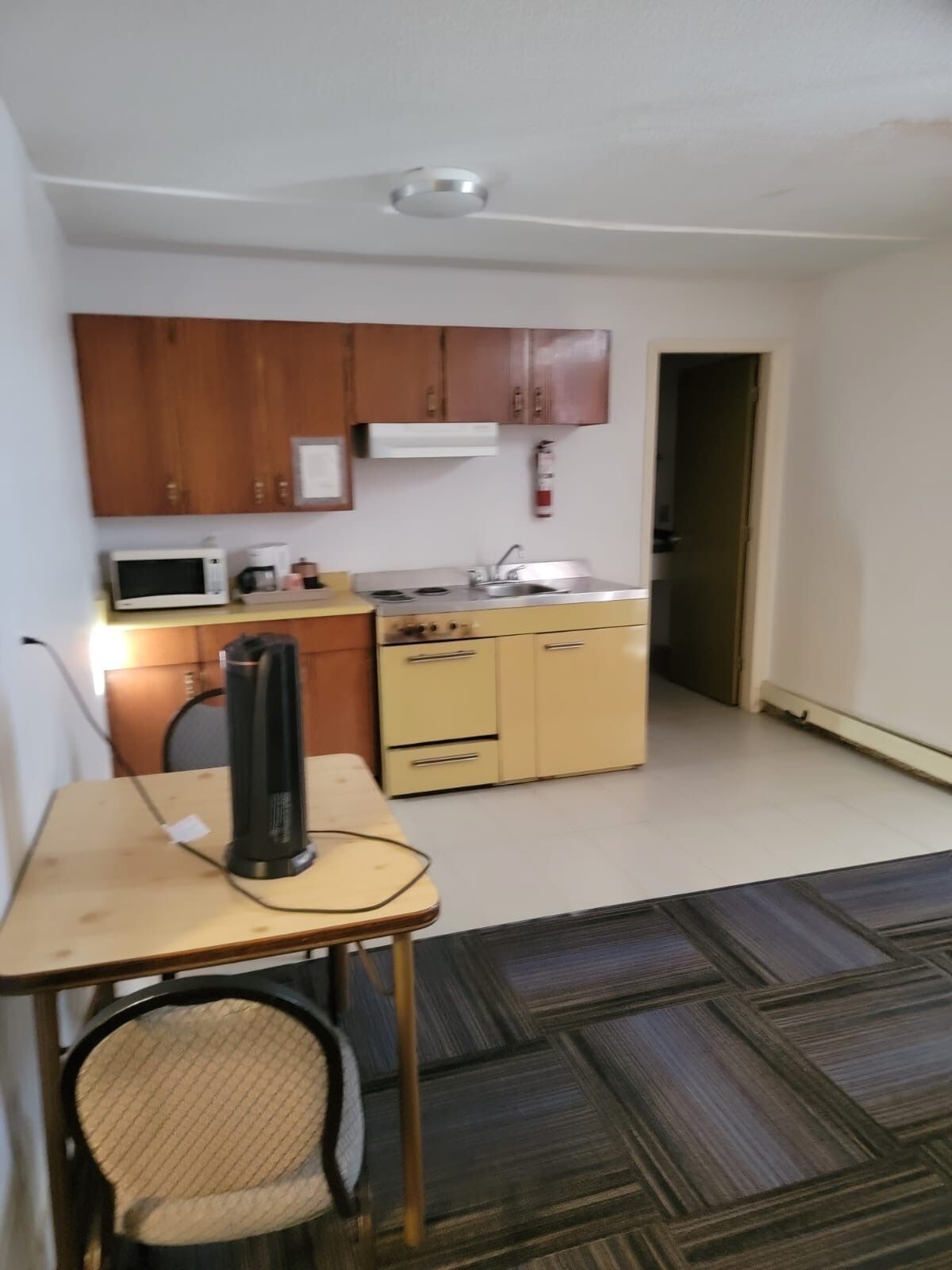 Cocina compacta con armarios de madera, microondas, estufa y lavaplatos. Hay una pequeña mesa con una silla y un objeto negro encima.