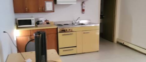 Cocina compacta con armarios de madera, microondas, estufa y lavaplatos. Hay una pequeña mesa con una silla y un objeto negro encima.