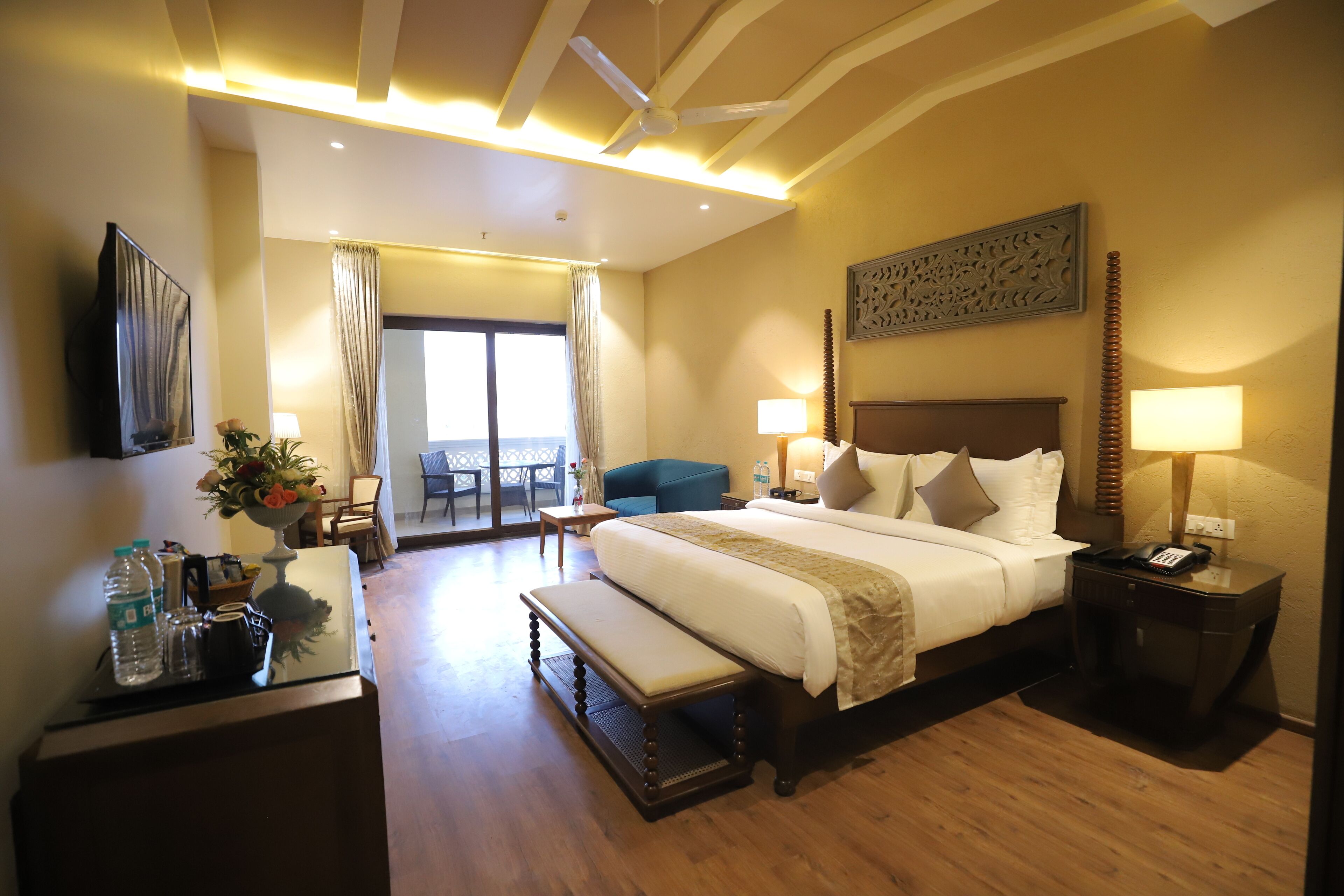 A hotel room with a large bed, bedside tables, a desk with a chair, a bench, a dining area with a table and chairs, and a view through a glass door.