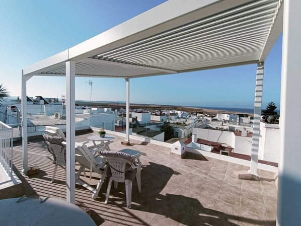 A rooftop terrace with a covered seating area, a view of the sea, and a cityscape in the background.