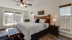 A bedroom with a bed, a bench, a bedside table, a lamp, a ceiling fan, and a window with a view of a backyard. - 3 King Bedrooms • Hot Tub • Game Room • Near Six Flags & SeaWorld (San Antonio)