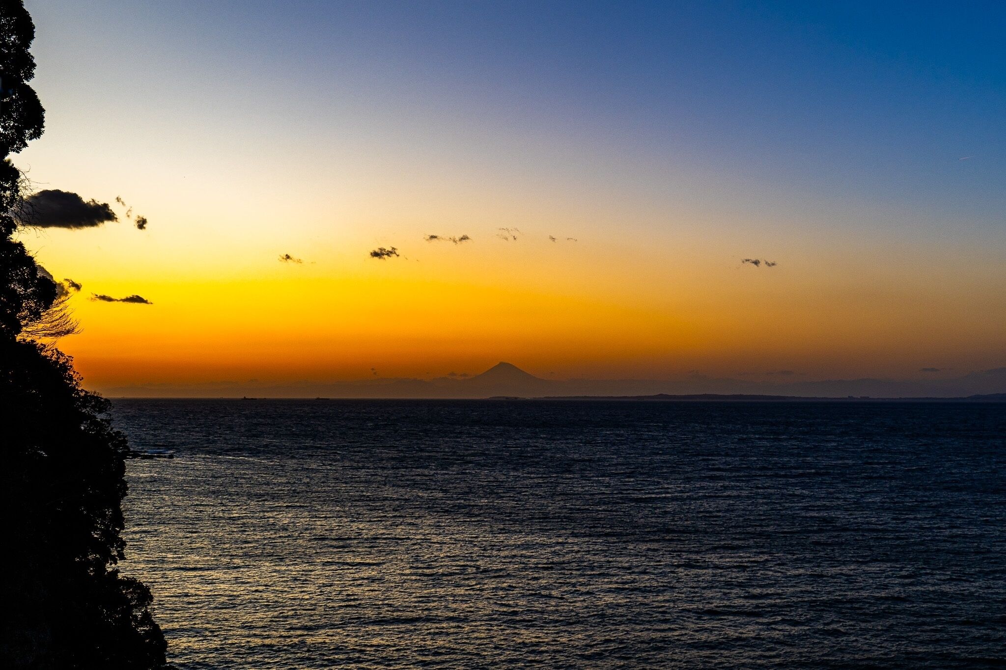 A sunset over a calm sea with a distant mountain and a rocky coastline.
