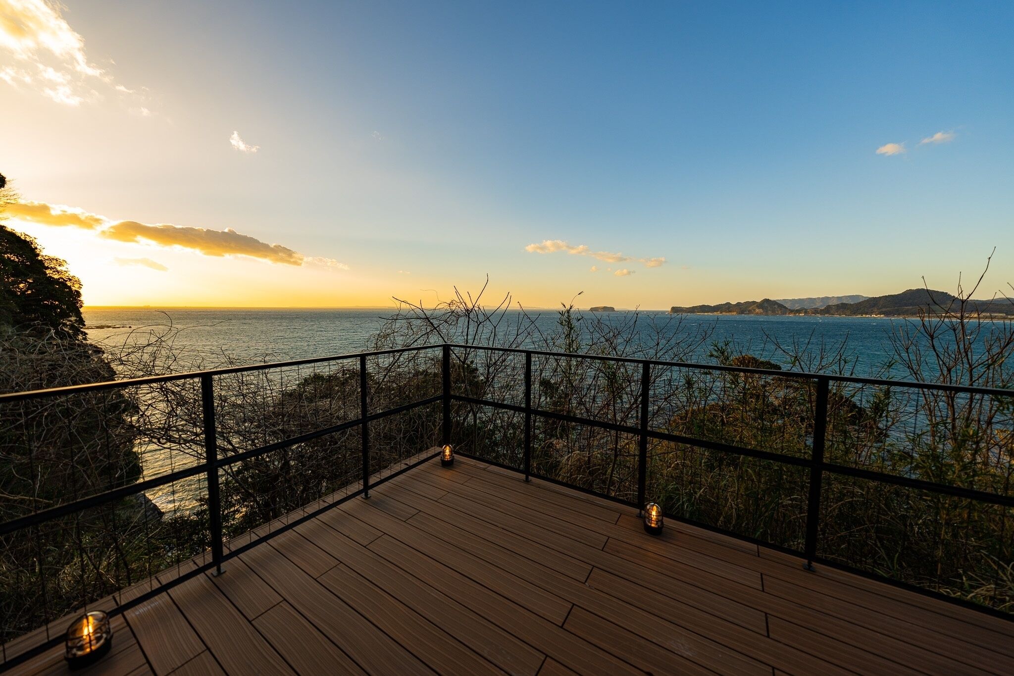 A wooden deck with a metal railing overlooking a coastal view during sunset.