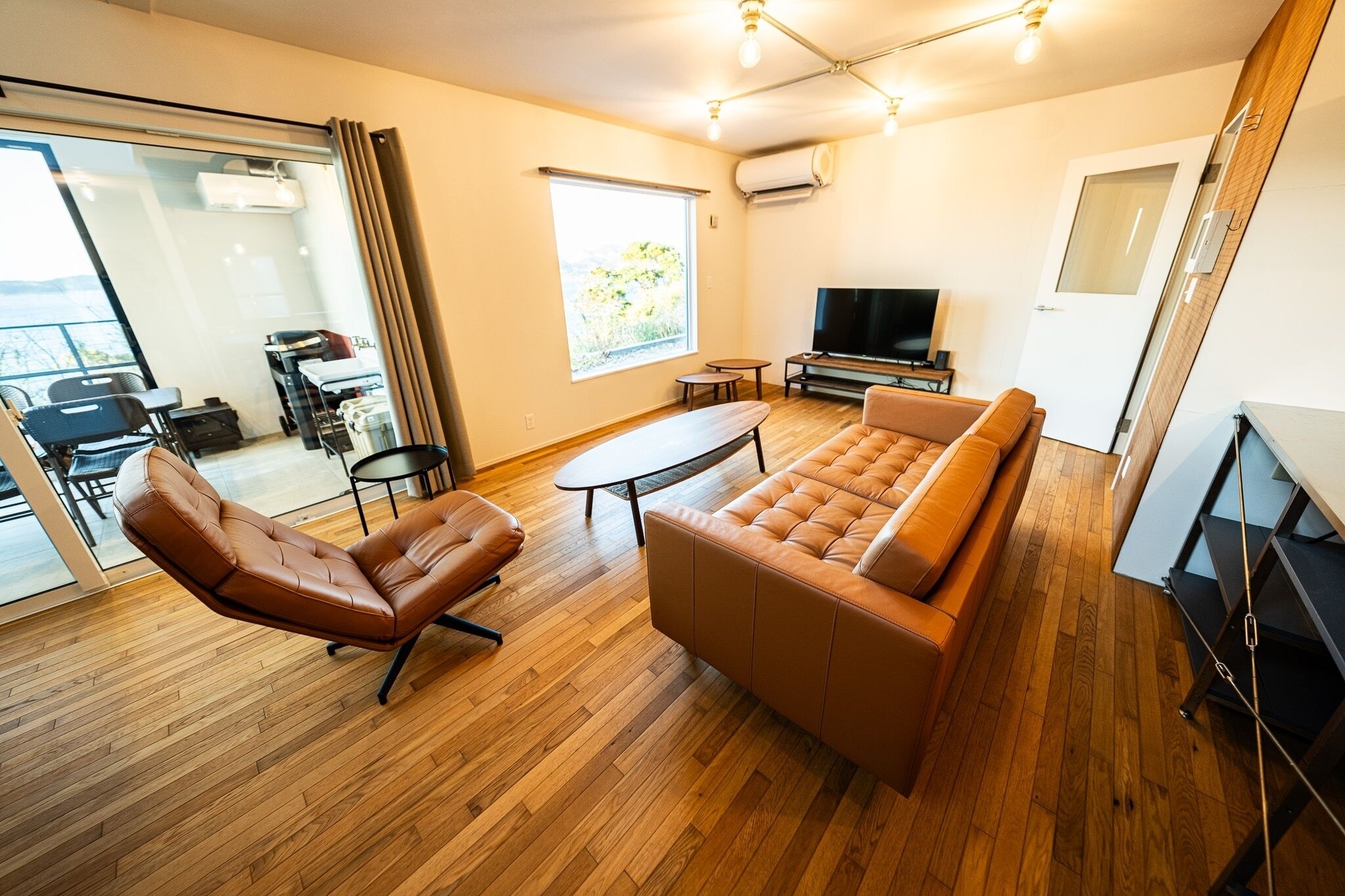 Una sala de estar moderna con un sofá, un sillón y una mesa de centro. Hay un televisor, una puerta corrediza a un balcón y una ventana con vistas.