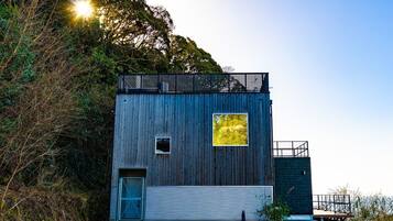 Eine moderne Holzhütte mit kleinem Fenster und Tür, umgeben von Bäumen und blauem Himmel.