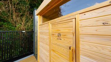 Una sauna in legno con una porta in vetro, un pontile di legno e una recinzione nera.