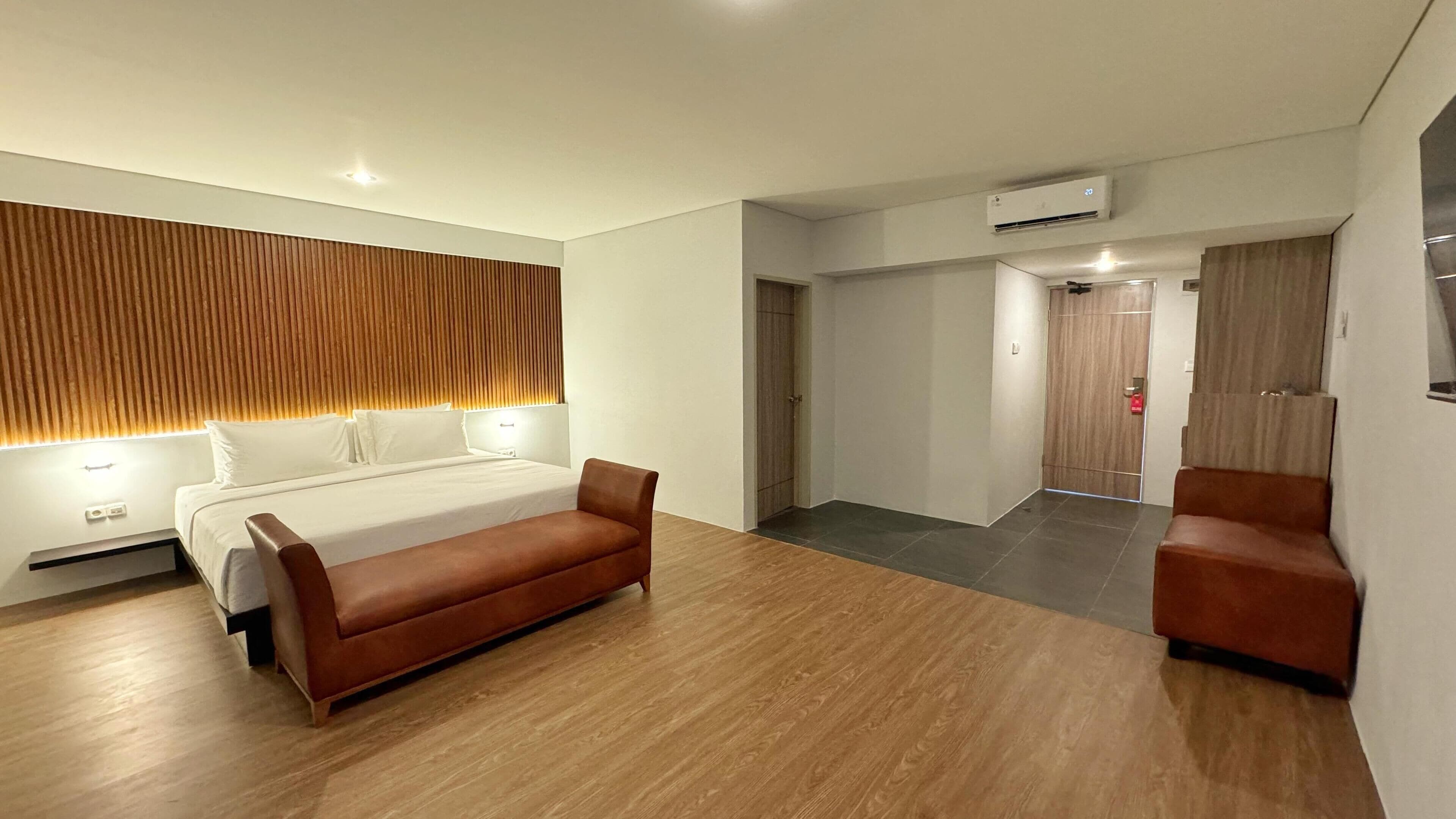 Una habitación de hotel moderna con una cama, un sofá marrón, suelo de madera y un equipo de aire acondicionado instalado en la pared.