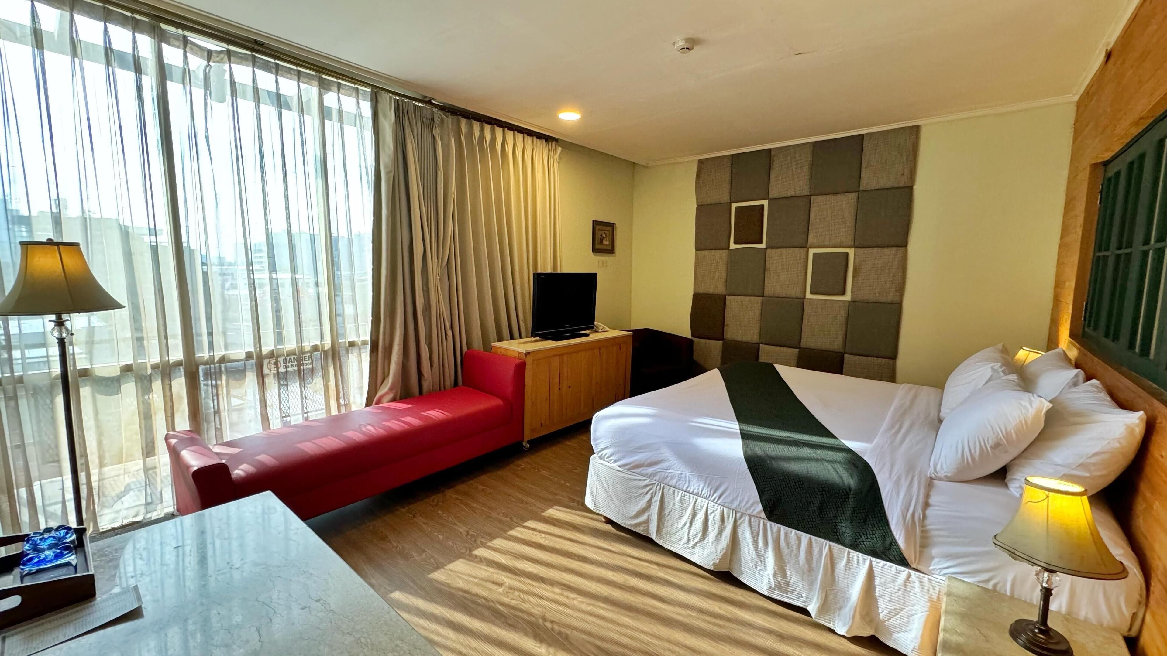 A hotel room with a bed, a red sofa, a wooden nightstand with a TV, a desk with a lamp, and a large window with curtains.