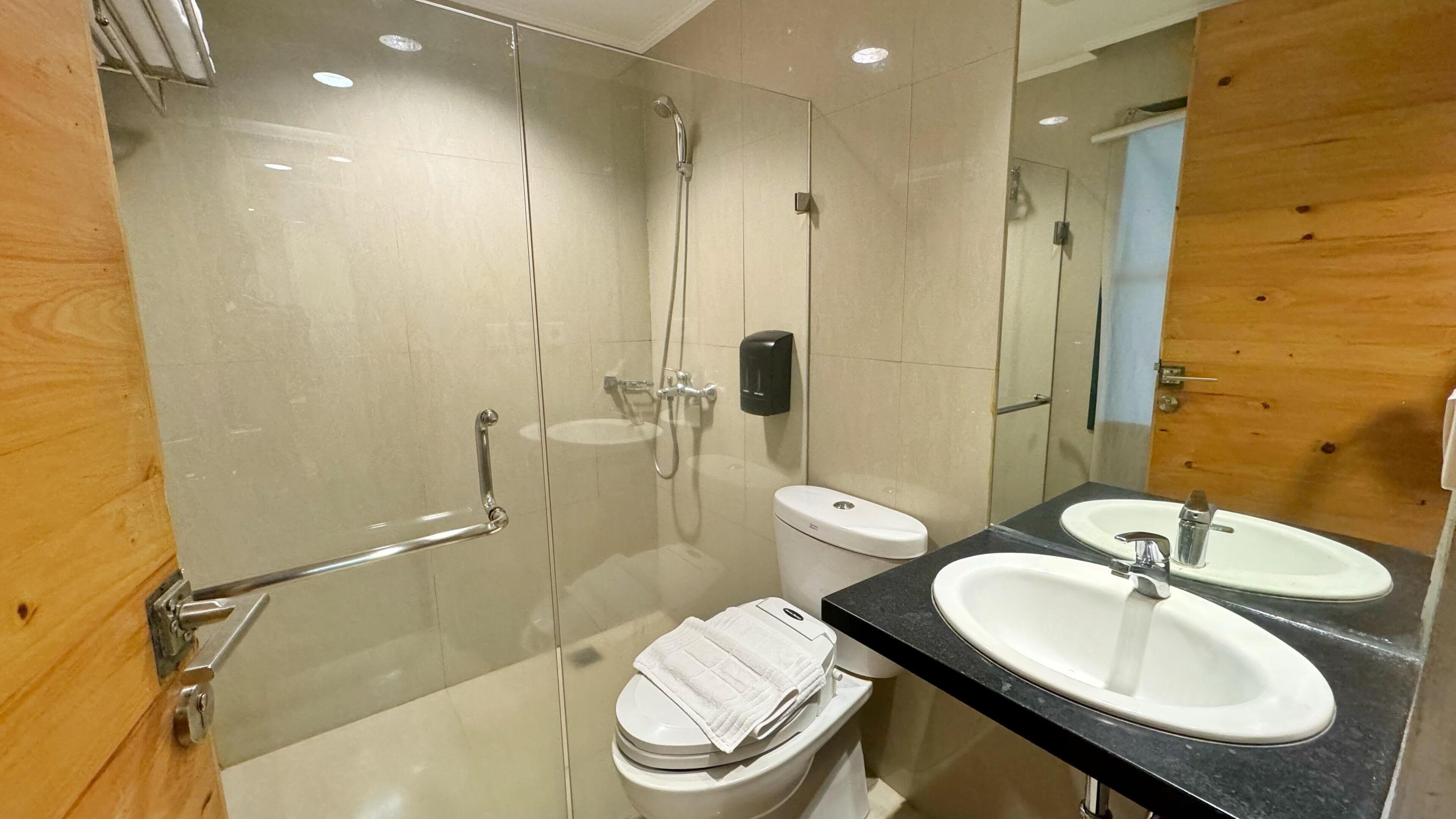 Een moderne badkamer met een douchecabine van glas, een toilet, twee wastafels en een houten wand.