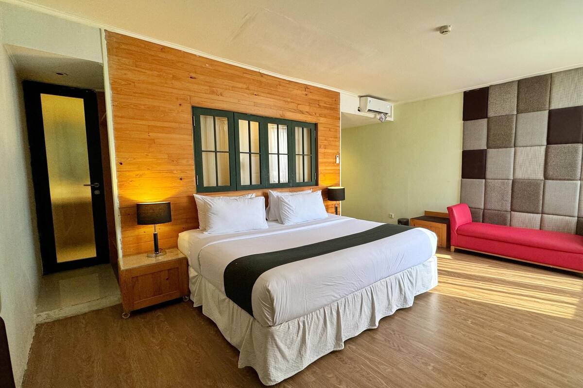 A hotel room with a wooden headboard, a bed with white bedding, a red sofa, and a wall-mounted TV.