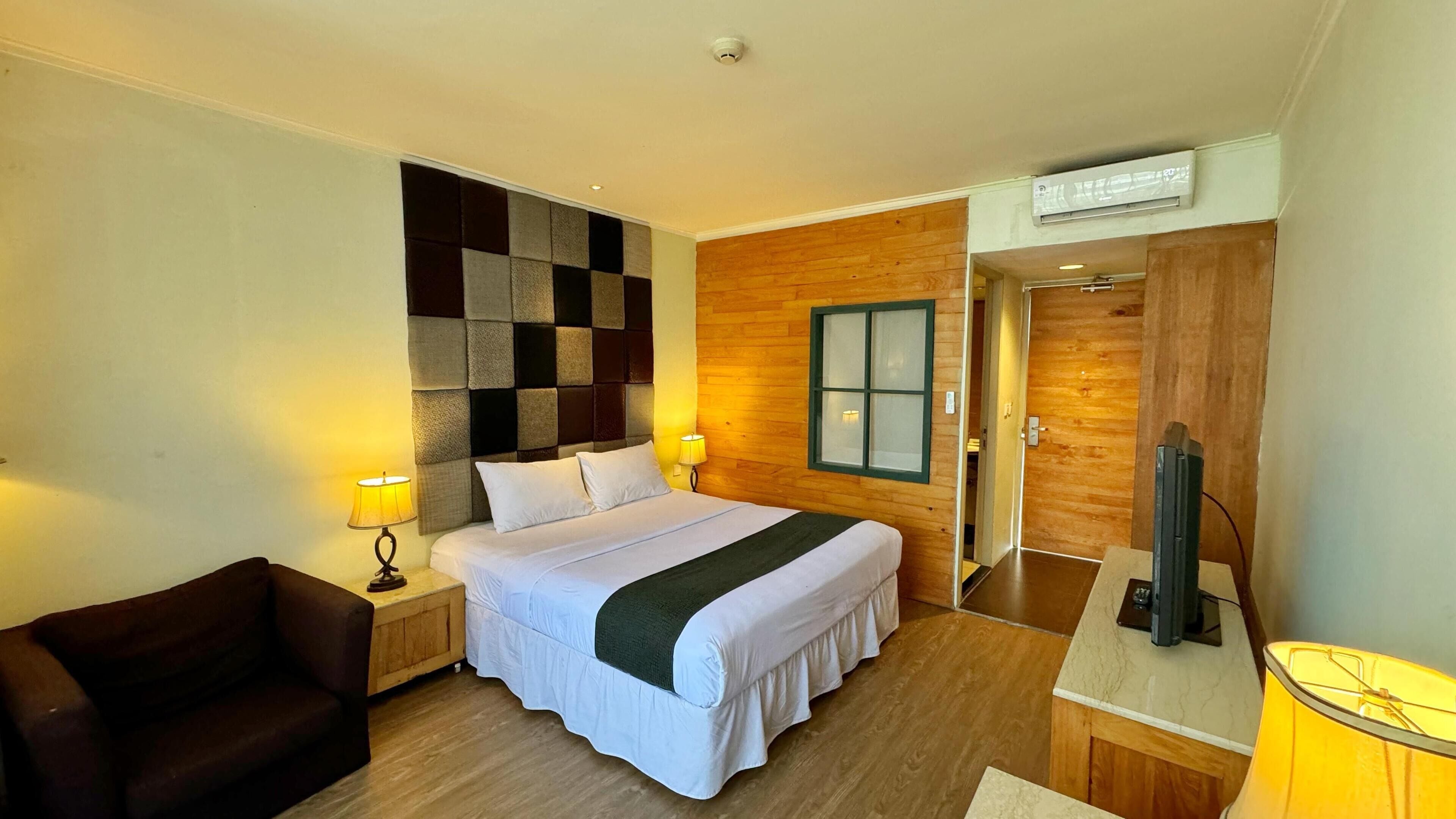 A hotel room with a bed, two bedside lamps, a wooden headboard, a window, a TV, a chair, and a wooden floor.