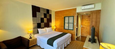 A hotel room with a bed, two bedside lamps, a wooden headboard, a window, a TV, a chair, and a wooden floor.