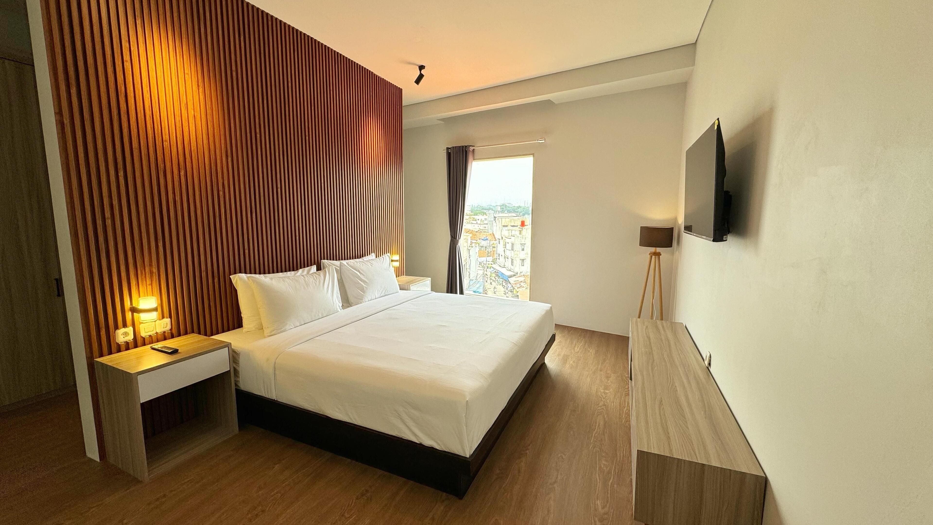 A modern hotel room with a large bed, a bedside table with a lamp, a TV mounted on the wall, and a view of the city through the window.