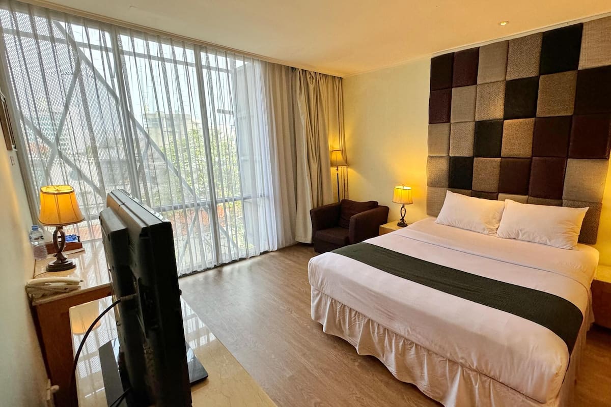 A hotel room with a large bed, a television, two side tables with lamps, and a view through a large window with sheer curtains.