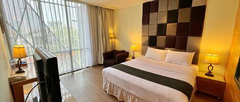 Habitación de hotel con una cama grande, un televisor, dos mesitas de noche con lámparas y vistas a través de un ventanal con cortinas translúcidas.