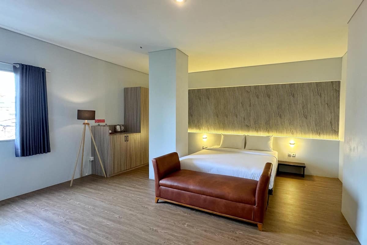 A modern hotel room with a large bed, a brown leather sofa, a desk with a lamp, and a view of the outdoors through a window with curtains.