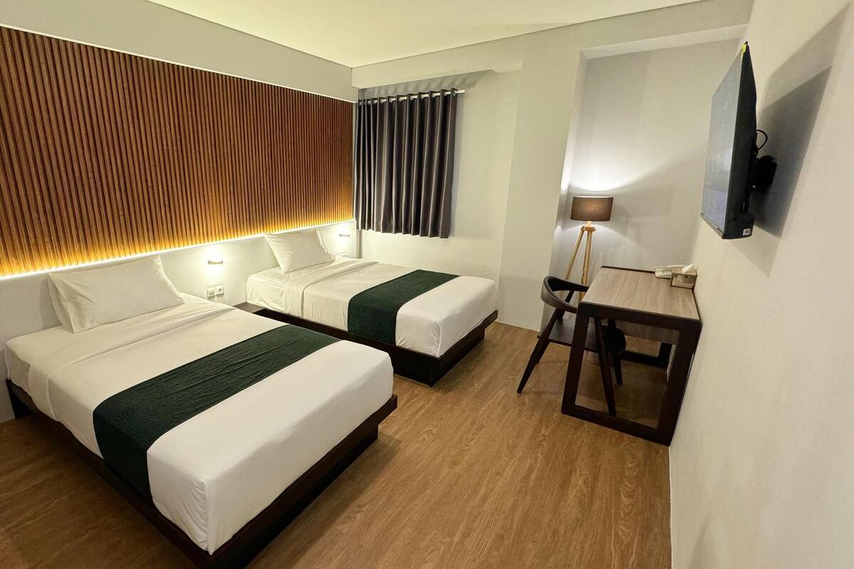 A hotel room with two beds, a desk, a chair, a TV, and a lamp.
