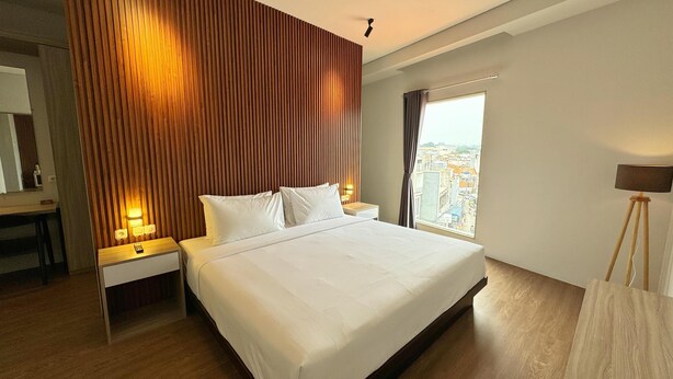 A hotel room with a large bed, wooden panel wall, bedside lamps, a desk, and a view of the city. - Chara Hotel (Bandung)