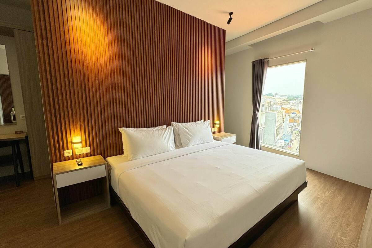 A hotel room with a large bed, wooden panel wall, bedside lamps, a desk, and a view of the city.