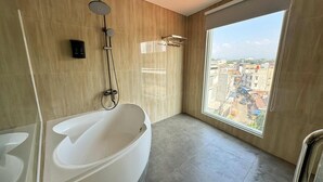 A modern bathroom with a large bathtub, a shower, and a toilet. There is a window providing a view of the city. - Chara Hotel (Bandung)