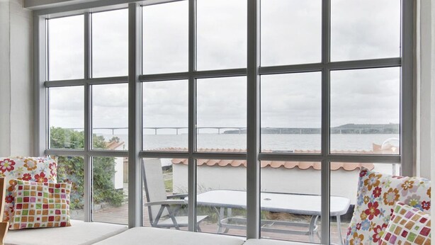 A room with a large window overlooking a body of water and a bridge. There is a bench with cushions and a table outside the window. - "Pauliina" - 10m to the fjord by Interhome (Roslev)
