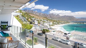 A balcony with a view of a coastal town, a road with cars, and a mountain range in the background. - Chic Apartment w Amazing Views Dunmore Luxe (Cape Town)