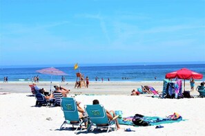 A beach with people relaxing on chairs and towels, umbrellas, and a lifeguard tower.