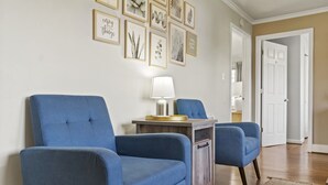 Olohuoneessa on kaksi sinistä nojatuolia, pieni puinen pöytä, jossa on lamppu, ja seinä, joka on koristeltu kehystetyillä taideteoksilla.