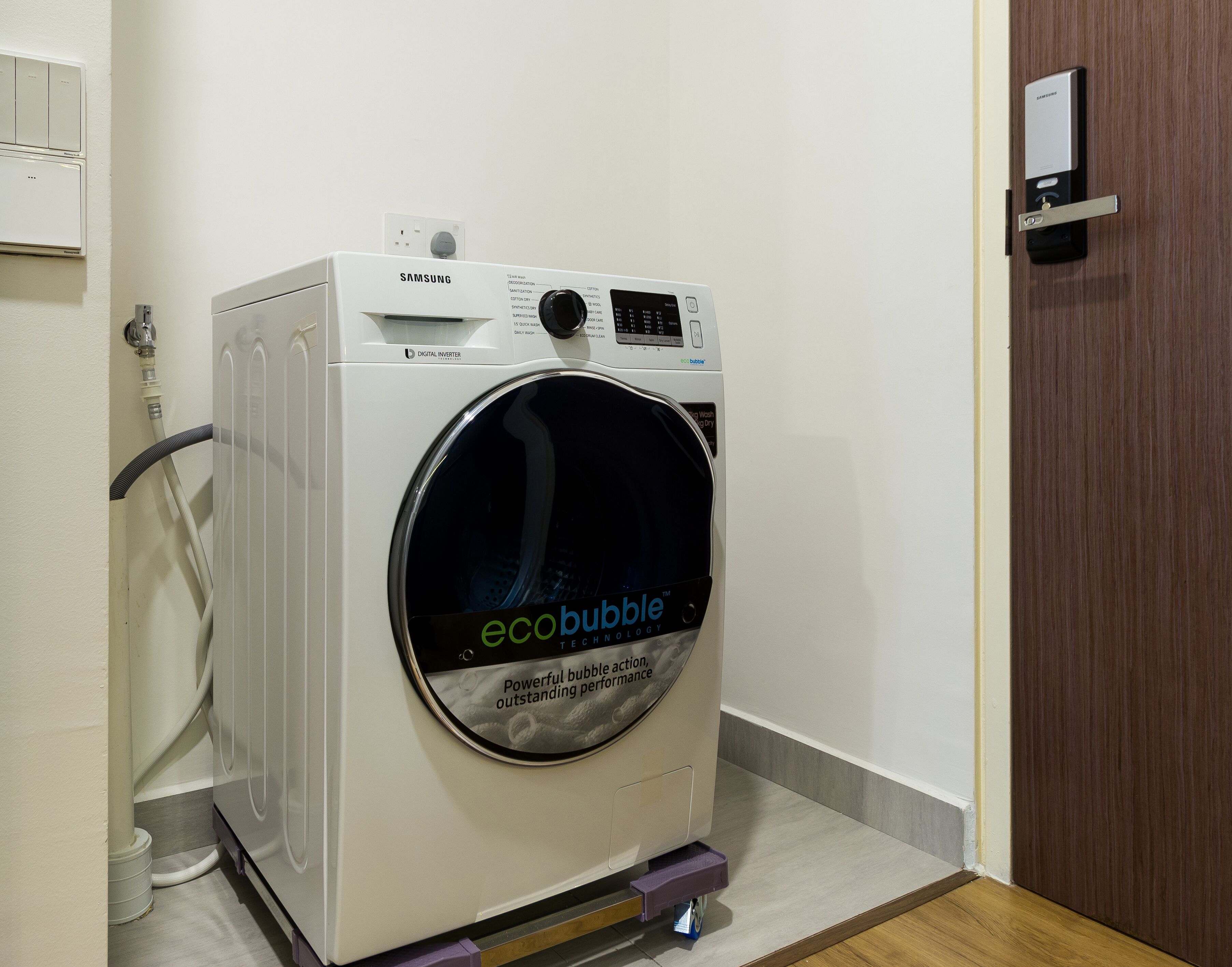 Una lavadora digital con tecnología Inverter Eco-Bubble de Samsung en una habitación con una puerta de madera y pared blanca.