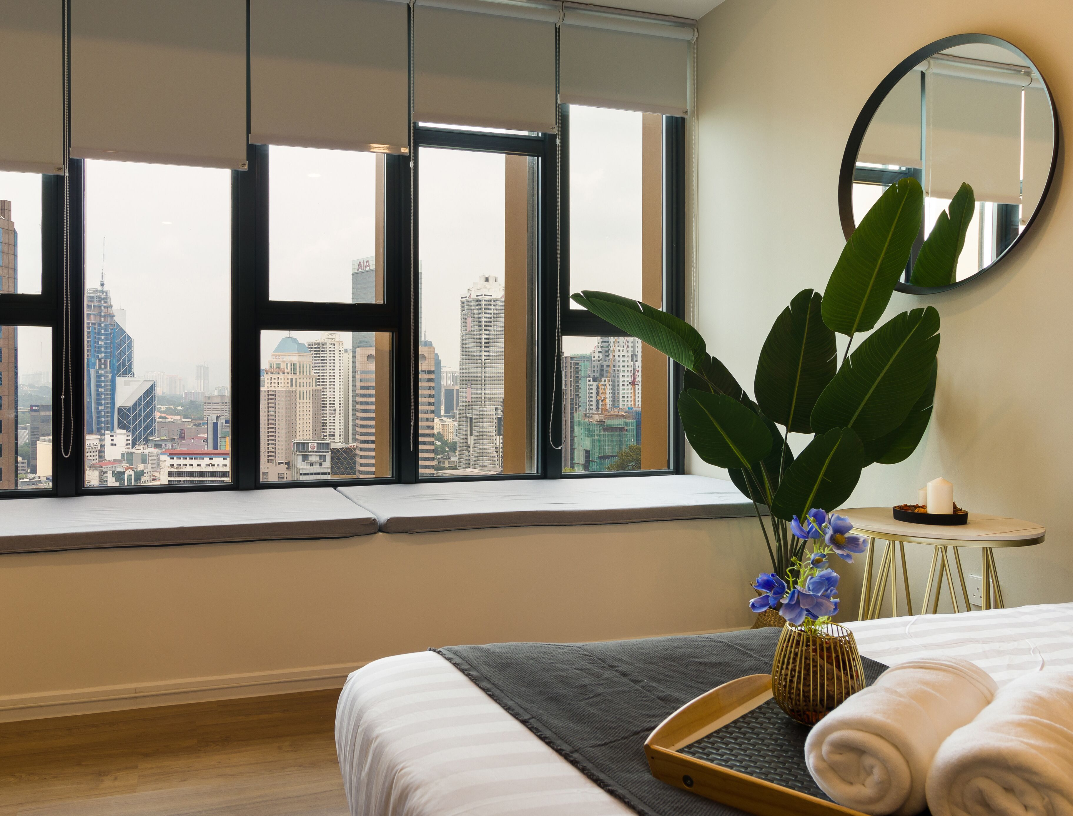 Quarto de hotel com uma janela grande que oferece vista para a cidade, uma cama com roupa de cama branca, uma mesa redonda pequena com uma vela e uma planta em vaso.