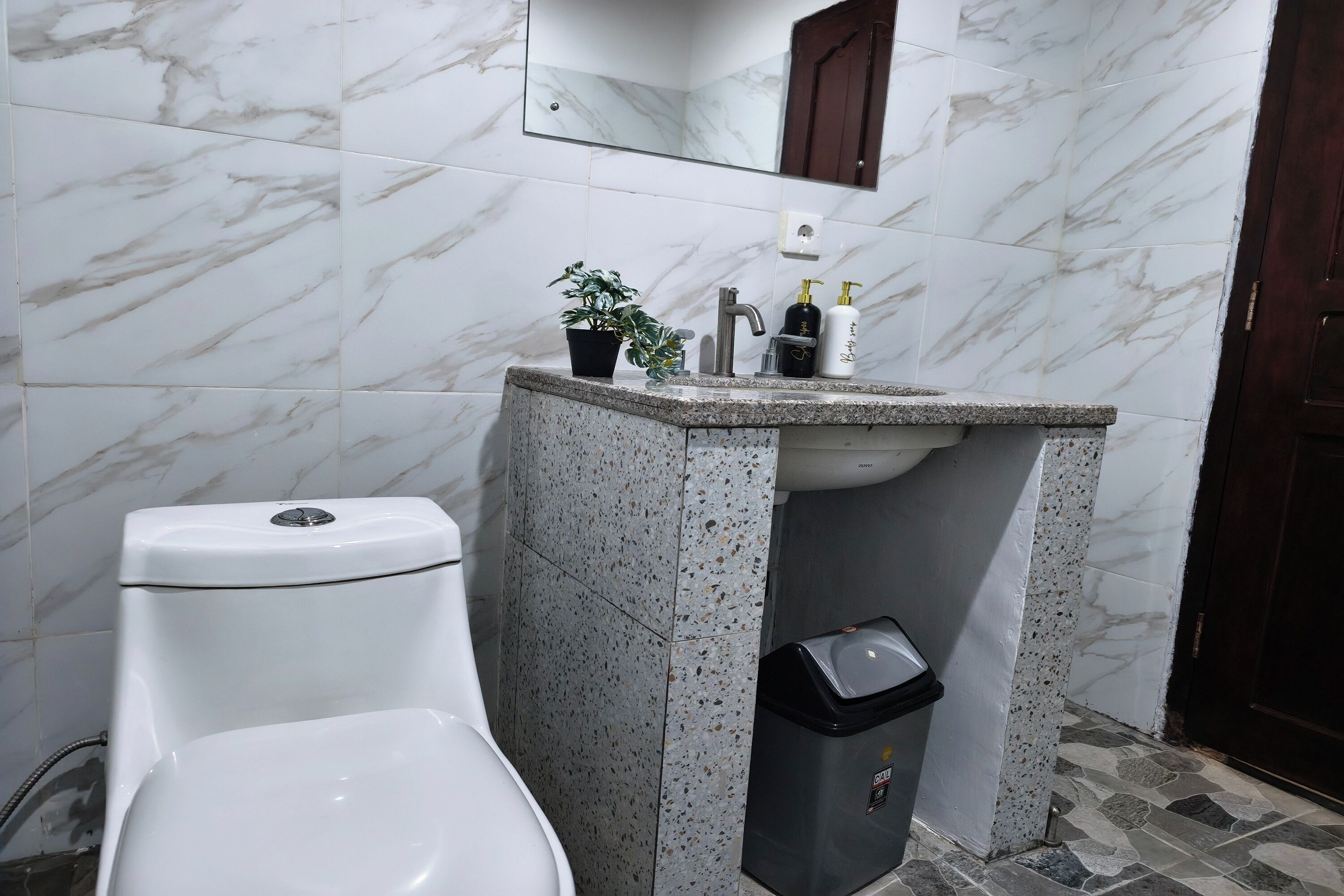 A bathroom with a toilet, sink, mirror, and a trash bin.