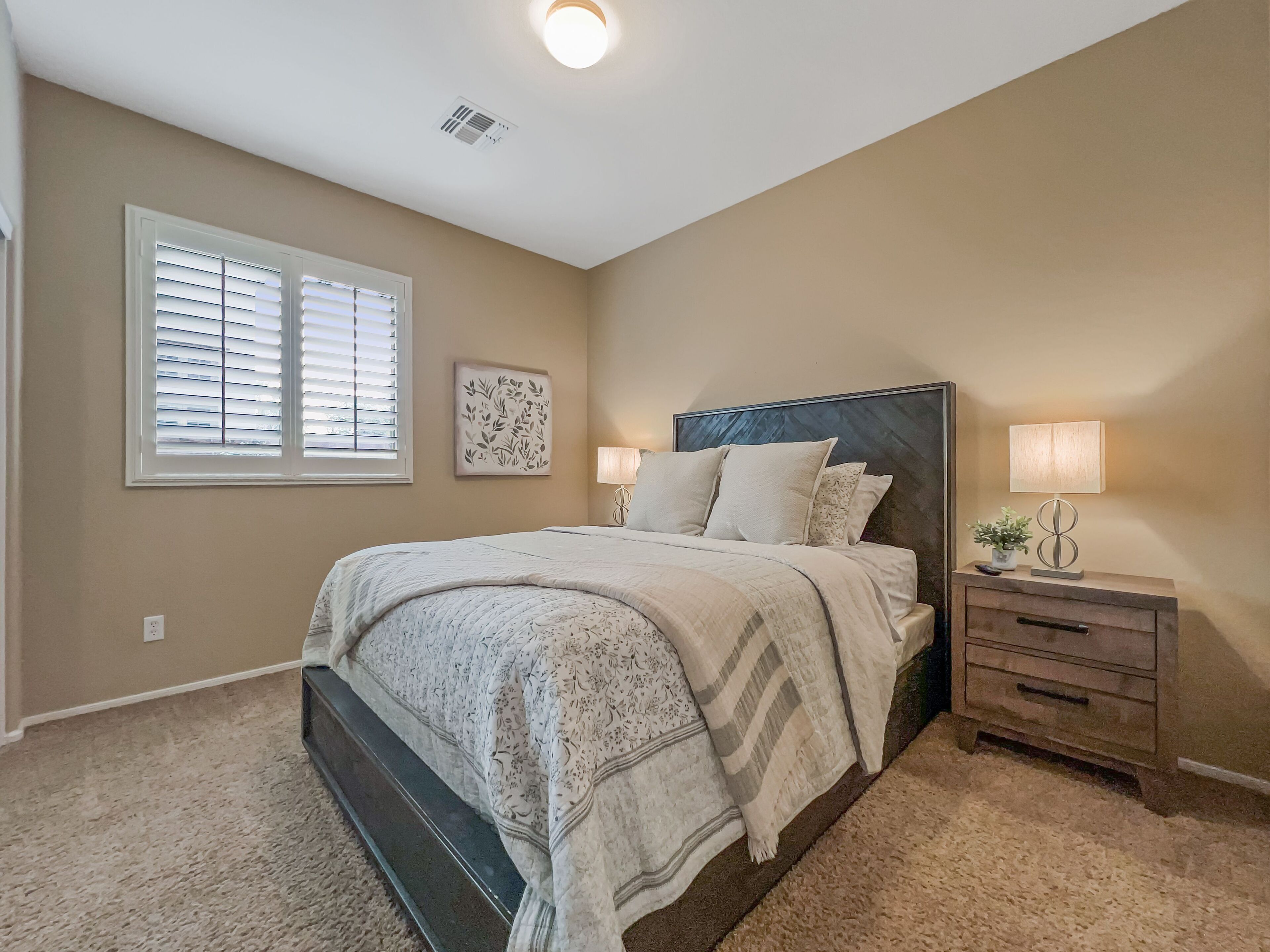 A bedroom with a bed, bedside tables, a window with blinds, a ceiling vent, and a wall-mounted artwork.