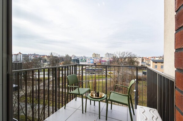 A balcony with two chairs and a table, overlooking a cityscape with buildings and a train track.