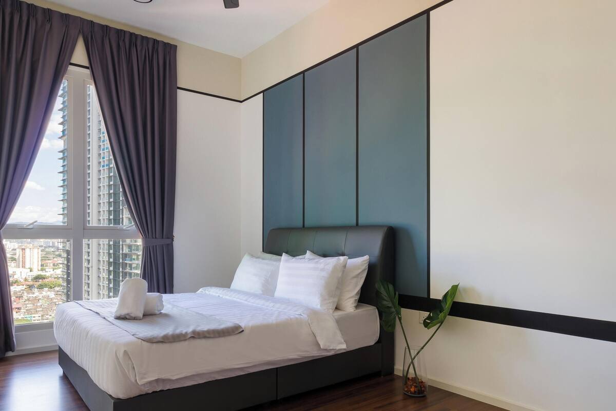 Ein modernes Schlafzimmer mit einem großen Bett, dunklen Vorhängen, Blick auf Gebäude und einer Zimmerpflanze.