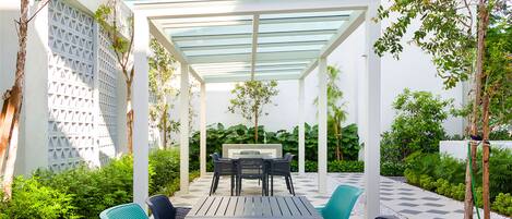 Un patio al aire libre con una mesa y sillas, rodeado de follaje y una pérgola con techo de vidrio.