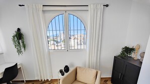 A room with a large arched window, a beige armchair, a black desk with a chair, a black cabinet, and a hanging plant. - AP393 LOS ALMENDROS (Nerja)