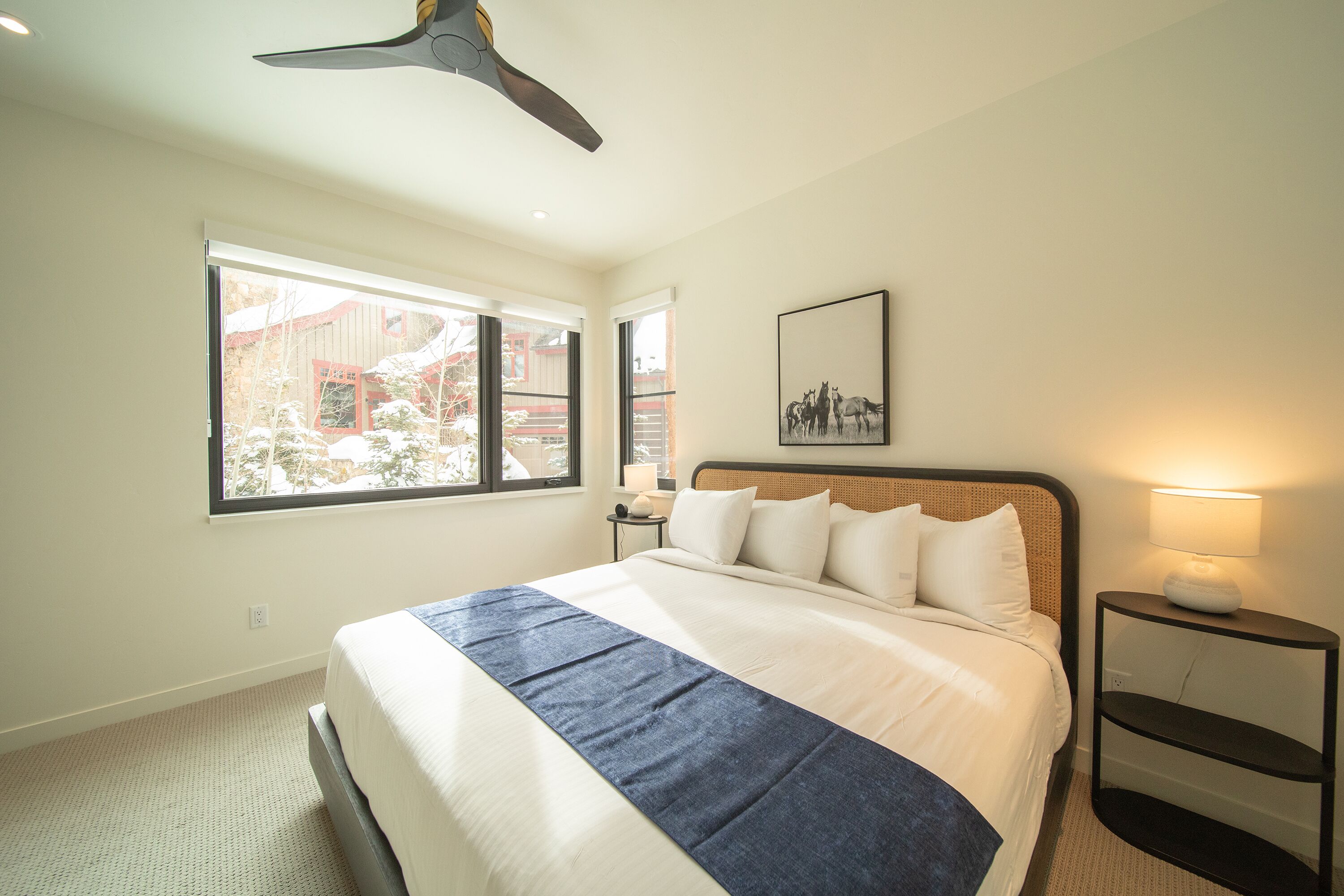 A hotel room with a large bed, a ceiling fan, a window with a view of buildings, a bedside table with a lamp, and a framed picture on the wall.