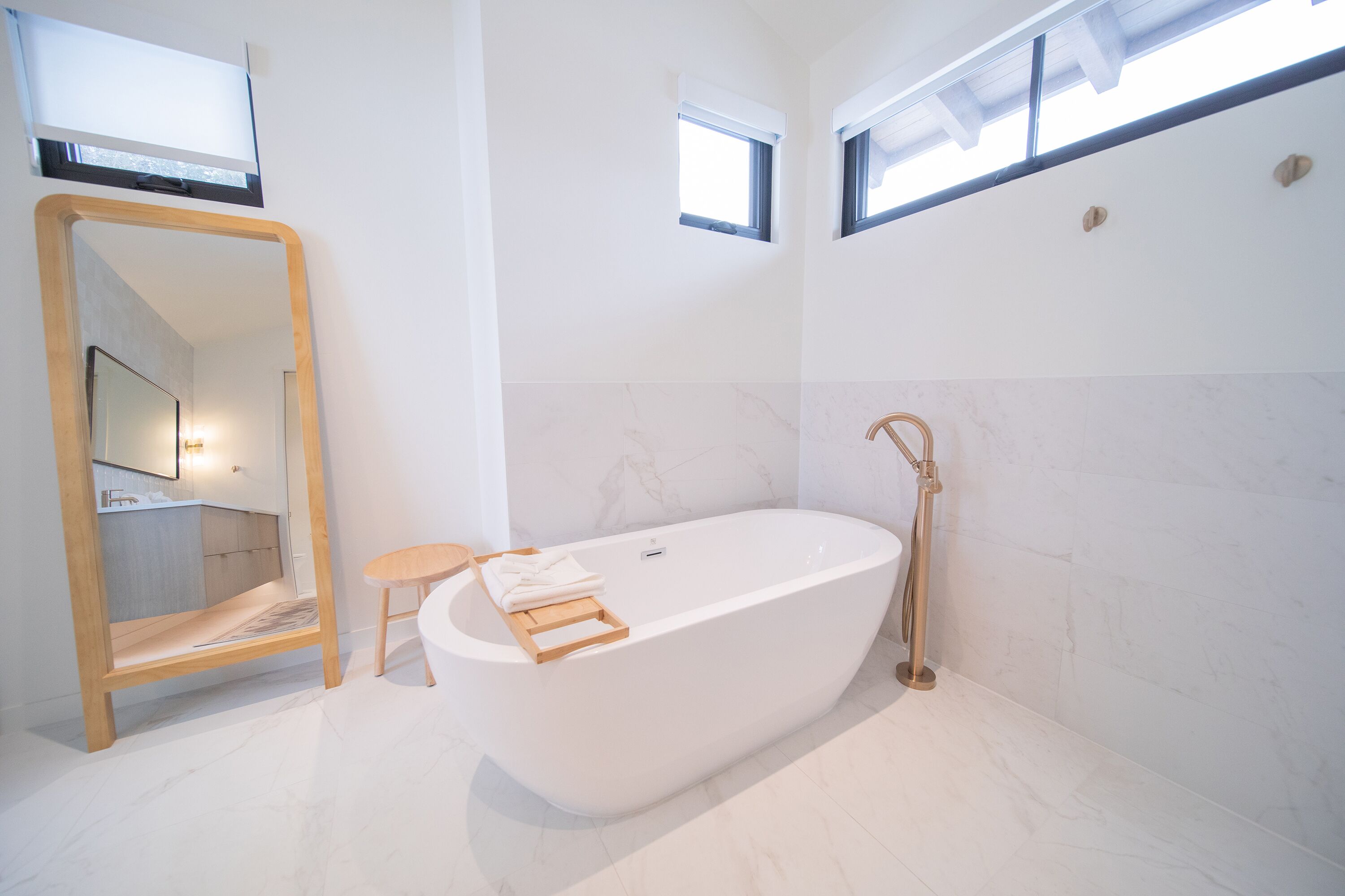 A modern bathroom with a freestanding bathtub, a wooden stool, a large mirror, and a window providing natural light.
