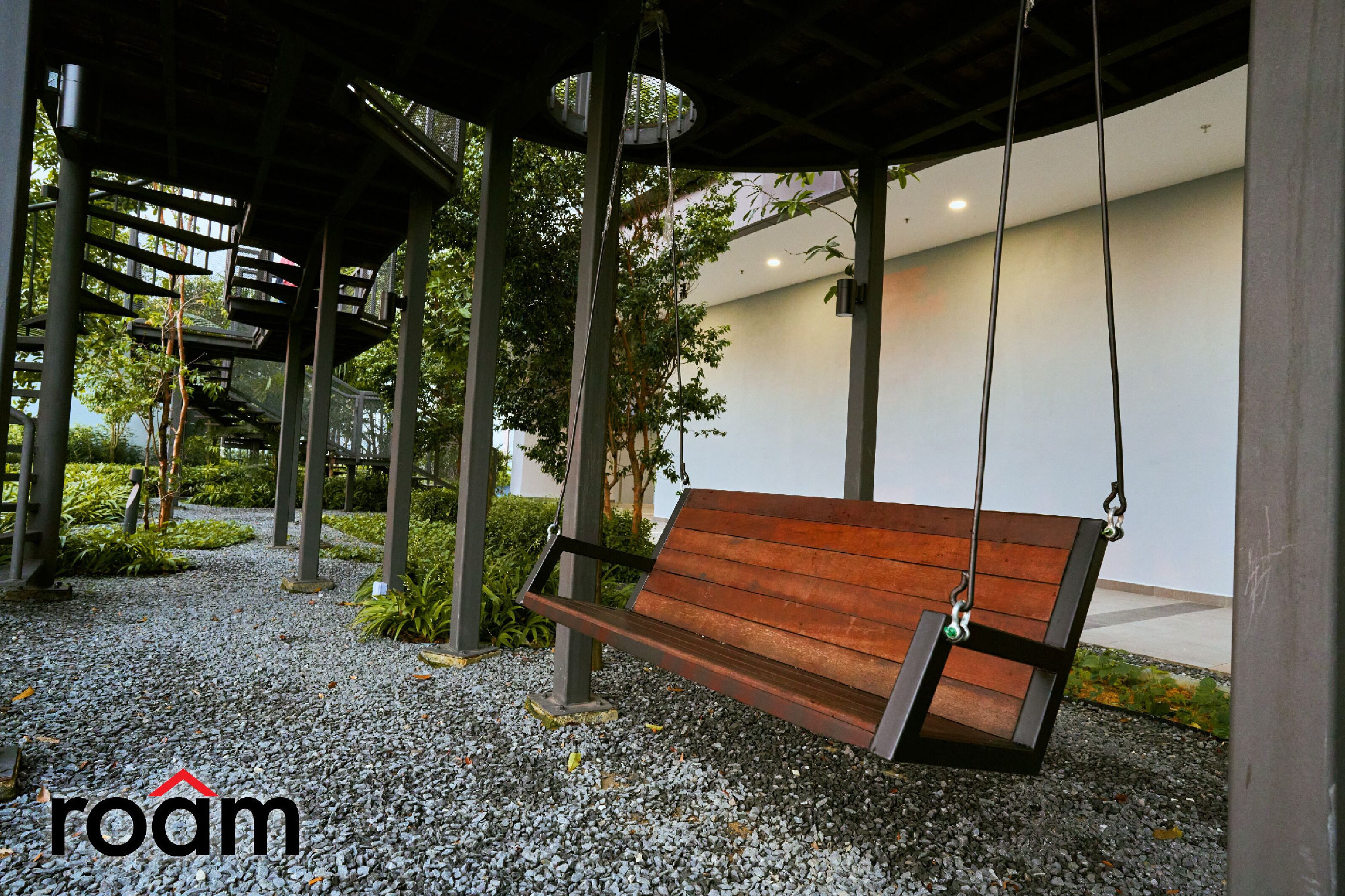 A wooden swing bench under a covered area with a gravel ground, surrounded by greenery and modern buildings.