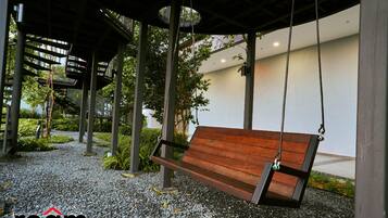 A wooden swing bench under a covered area with a gravel ground, surrounded by greenery and modern buildings.
