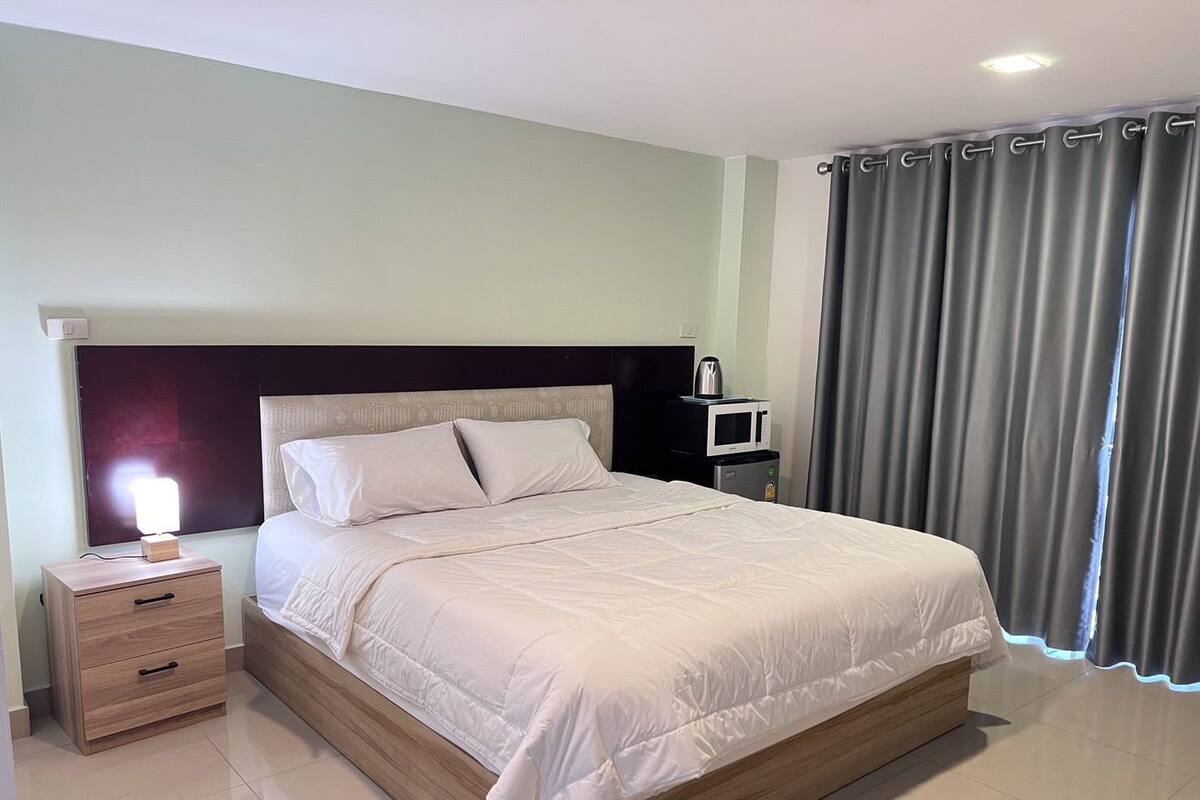 Una cama bien hecha con cabecera de madera, una mesita de noche con lámpara y un microondas sobre una pequeña mesa al lado de la cama.