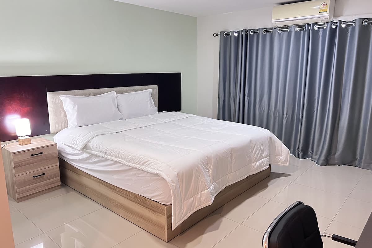 Una habitación de hotel con una cama, una mesita de noche con lámpara, una silla y un ventanal con cortinas.