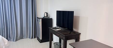 Ett kompakt hotellrum med en säng, ett skrivbord med en dator, ett litet kylskåp och en gardin.