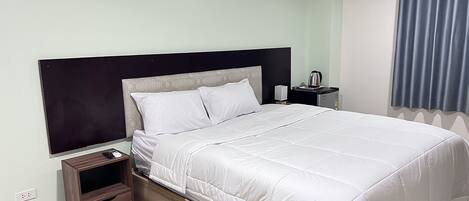 A neatly made bed with white bedding, a wooden nightstand with a drawer, a small table with a kettle, and a window with blue curtains.